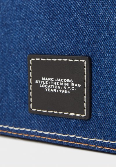 Geantă mini din denim cu un material texturat de culoare albastră, având un eticheta neagră conturată cu cusături albe care include detalii de brand și design.