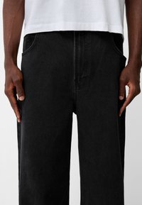Pantalones de mezclilla negros con un corte relajado, que cuentan con bolsillos laterales, un sutil detalle de costura y una tela texturizada, combinados con una camiseta blanca.