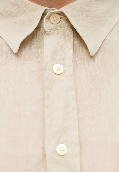 Μπεζ λινό πουκάμισο με γιακά που κουμπώνει με κουμπί, διαθέτει δύο λευκά κουμπιά και υφή υφάσματος. Απλός, κλασικός σχεδιασμός με φυσική εμφάνιση.
