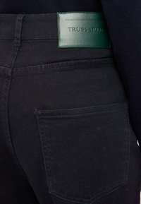 El bolsillo trasero de unos jeans de mezclilla oscura con una etiqueta de cuero verde cosida encima del bolsillo que muestra el nombre de la marca "TRUSSARDI".
