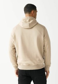 Beige hættepullover lavet af blødt materiale, med en kengurulomme og ribbede manchetter, parret med sorte bukser.