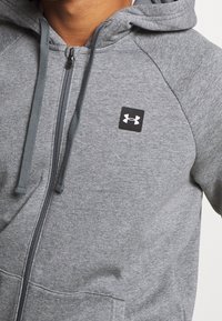 Felpa grigia con zip realizzata in tessuto morbido. Presenta un cappuccio, stringhe regolabili e un logo quadrato nero sul petto. Include una tasca frontale.