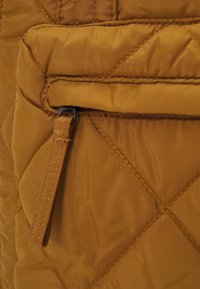 Tecido acolchoado em amarelo mostarda com uma costura vertical e um pequeno bolso com zíper e puxador metálico. Textura suave.