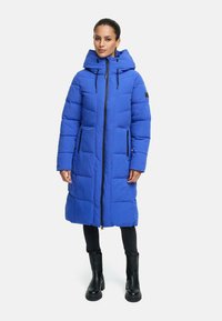 Heldblau pufferjack met capuchon, met een volledige ritssluiting aan de voorkant, zijzakken en een doorgestikt ontwerp. Gepaard met zwarte leggings en laarzen.