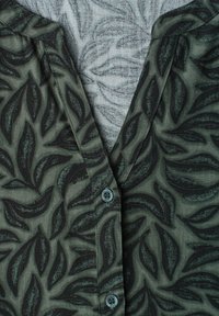 Chemise vert foncé avec des motifs de feuilles noires, partiellement boutonnée, laissant apparaître une chemise gris clair avec des motifs abstraits de feuilles noires en dessous.