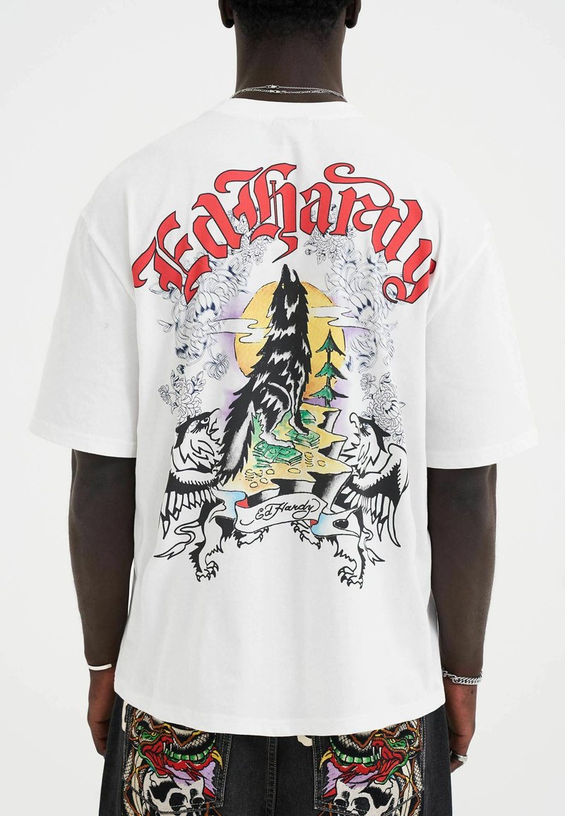 Hvid t-shirt med farverigt grafik af en ulv, træer og fugle med teksten "Ed Hardy", designet med intrikate linjer og detaljer.