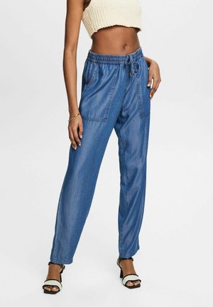 Frau trägt hoch taillierte blaue Jeans-Jogginghosen mit Vordertaschen, kombiniert mit weißem Strick-Crop-Top und schwarz-weißen Sandalen mit Absatz.