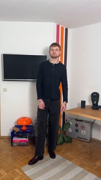 Black long-sleeve henley shirt, dark trousers, brown shoes, standing in a room with a TV, orange lamp, and a wood table with accessories.