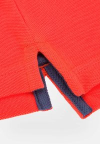 Czerwony dzianinowy element odzieży z granatowymi akcentami, posiadający rozcięty dół dla swobody ruchów oraz teksturowany materiał.