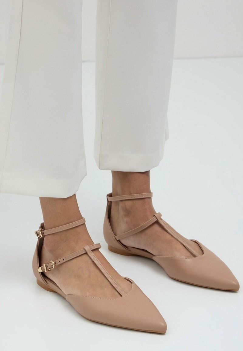 Pieds vêtus de ballerines beiges à bout pointu avec double lanières fines et boucles à la cheville, assortis à un pantalon blanc court sur un fond uni.