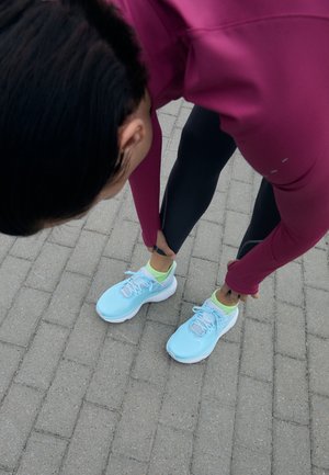 lichtblauwe hardloopschoenen van meshmateriaal met witte zolen, gecombineerd met zwarte legging en een bordeauxrood topje. Verhard oppervlak op de achtergrond.