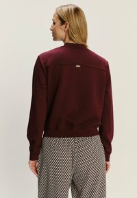 Bordeaux sweatshirt met een gestructureerde afwerking, voorzien van een ronde halslijn en een klein metalen accent op de rug, gecombineerd met gebloemde broek.