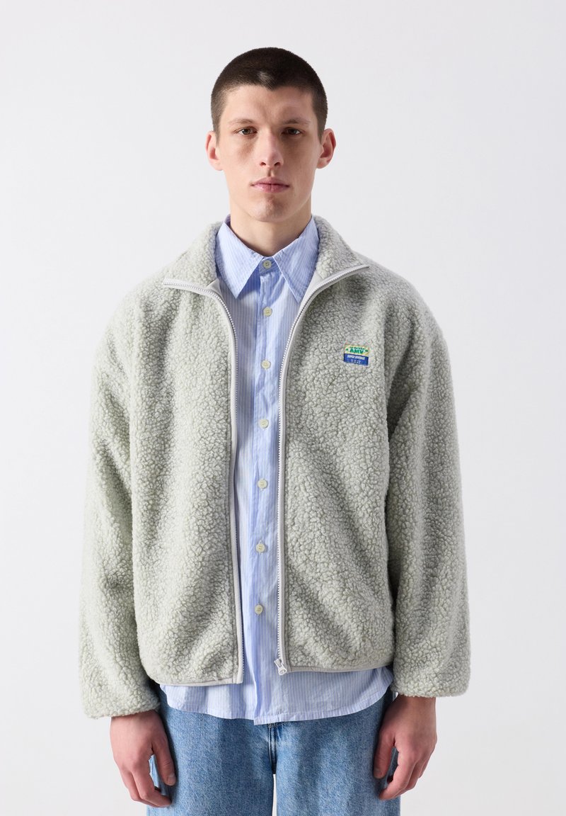 Mladý muž s krátkými vlasy, oblečený v světle modré košili na knoflíky, šedé fleecové bundě na zip a modrých džínách, stojící před jednobarevným pozadím.