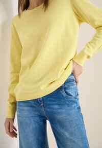 Femme portant un pull à manches longues jaune clair et un jean bleu, une main dans la poche, sur un fond neutre.