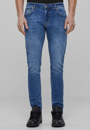 Blå denimjeans med slim fit, med falming og whiskering detaljer. Kombinert med svarte treningssko.