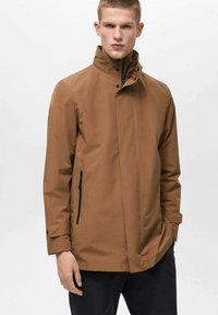 Brun vandtæt jakke med høj krave, trykknapper, sidelommer med lynlås og løs pasform; glat tekstur og minimalistisk design.