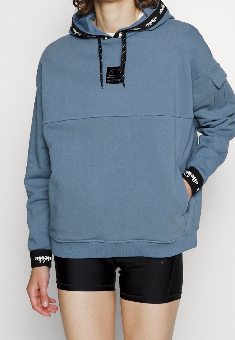 Personne portant un sweat à capuche bleu oversize avec des détails de logo noirs et un short de cyclisme noir, une main dans la poche, sur un fond uni.
