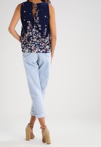 Mörkblå ärmlös topp med blommönster och bindning på baksidan, kombinerad med ljusblå jeans med uppvikta ärmar och beige högklackade sandaler.