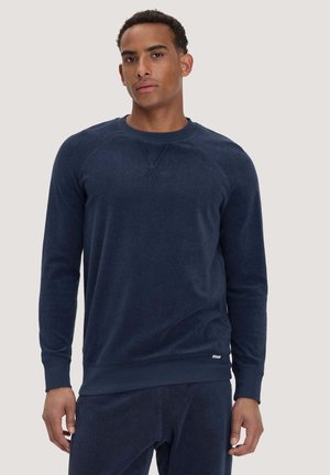 Mann trägt einen dunkelblauen Langarm-Sweatshirt und passende Hose, steht vor einem schlichten hellen Hintergrund.
