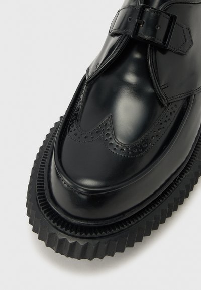 Μαύρο δερμάτινο παπούτσι με διακοσμητικές τρύπες (brogue), λουράκι με αγκράφα και παχύ, ραβδωτό πέλμα, που εμφανίζει το μπροστινό και το πλαϊνό άνω μέρος.