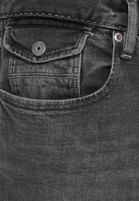 Jeans de mezclilla gris que cuentan con un pequeño bolsillo frontal con un cierre a presión metálico. La textura es suave con un desvanecimiento sutil y detalles de costura.