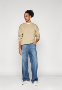 Sweat-shirt beige à col rond, logo sur la poitrine ; jean en denim bleu ample, motif délavé, baskets blanches avec trois bandes sur les côtés.