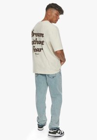 Beige kortærmet t-shirt med "Drøm uden frygt" trykt i brunt på ryggen, parret med lyseblå jeans og sorte Nike sneakers.