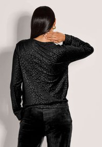 Vrouw met lang donker haar, gekleed in een zwart fluwelen tijgerprint top en bijpassende broek, hand op haar hals, met haar gezicht naar weg gericht.