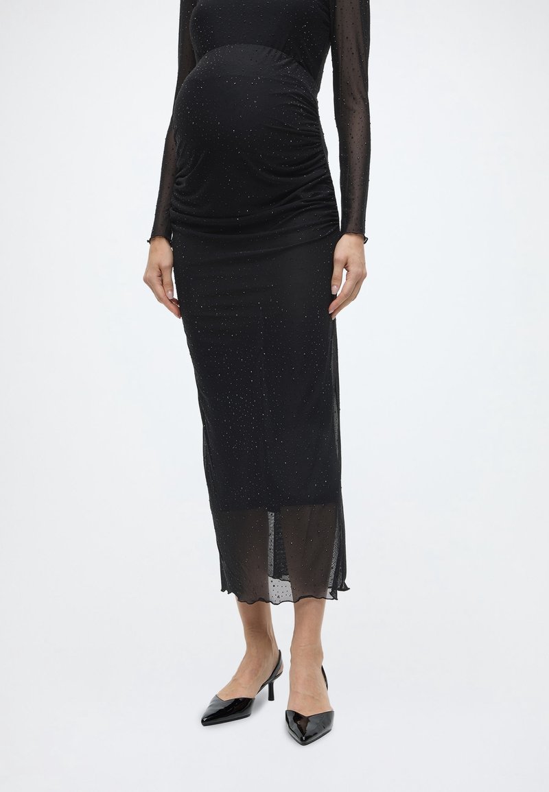 Femme enceinte portant une robe noire moulante à manches longues, scintillante, avec un tissu transparent en bas et des talons noirs à bout pointu.