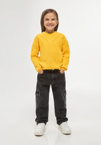 Maglione giallo a maglia con un motivo testurizzato, abbinato a pantaloni cargo neri e sneakers bianche. Il bambino sta in piedi con le mani nelle tasche.