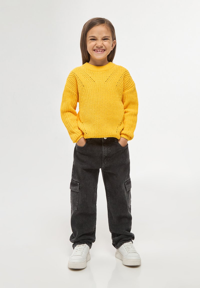 Maglione giallo a maglia con un motivo testurizzato, abbinato a pantaloni cargo neri e sneakers bianche. Il bambino sta in piedi con le mani nelle tasche.