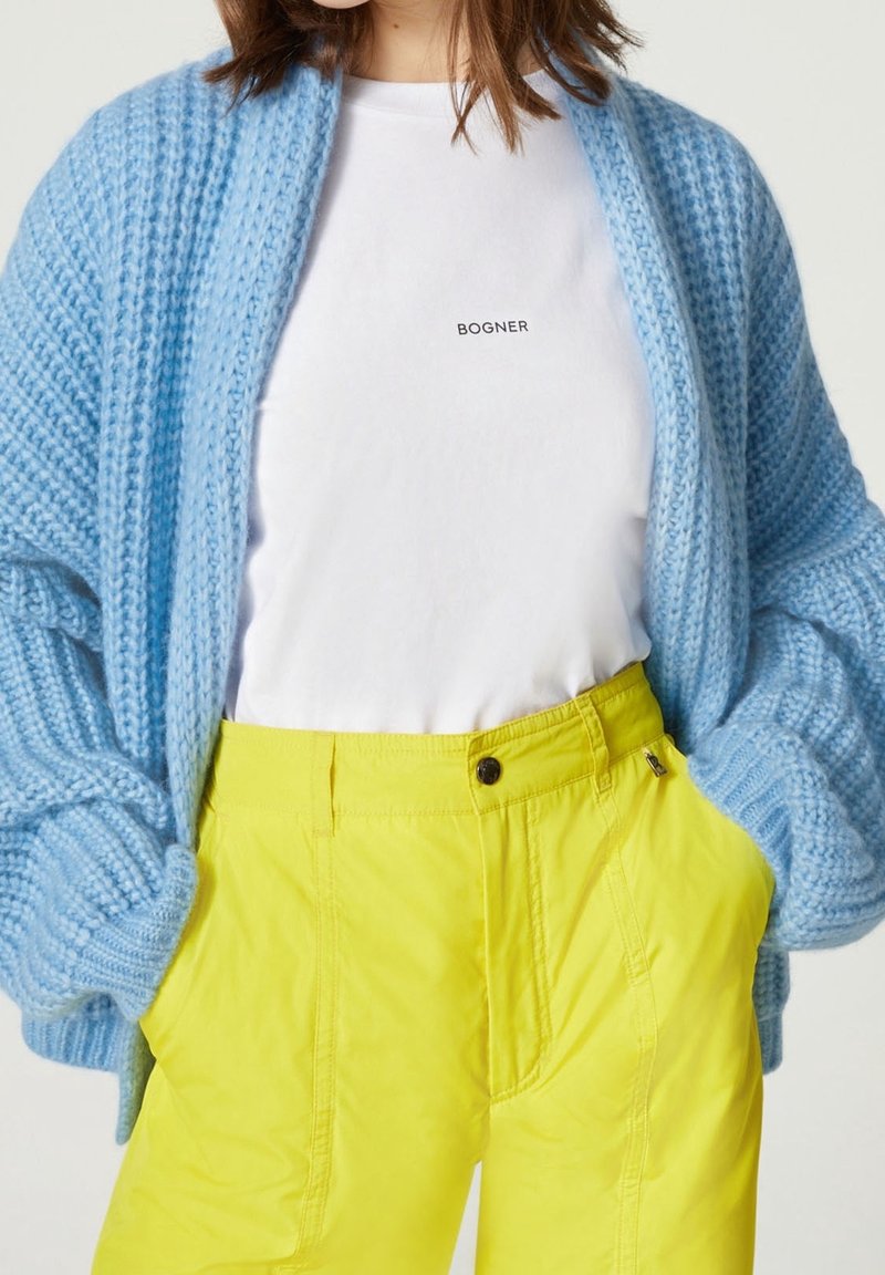 Personne portant un t-shirt blanc Bogner, un cardigan en grosse maille bleu clair, et un pantalon taille haute jaune vif, les mains dans les poches.