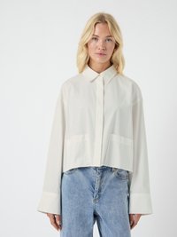 Blonde woman wearing a loose-fit, long-sleeve white shirt with large cuffs and light blue high-waisted jeans against white background.