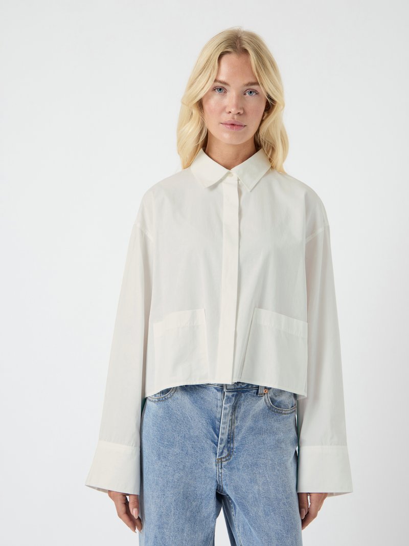 Ξανθιά γυναίκα που φοράει ένα χαλαρό, μακρυμάνικ ο άσπρο πουκάμισο με μεγάλα μανίκια και ανοιχτά μπλε ψηλόμεσα τζιν μπροστά σε λευκό φόντο.