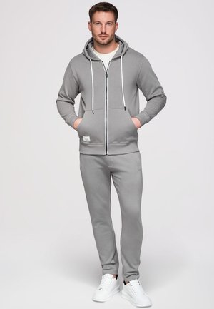 Homme debout face à l'avant portant un sweat à capuche gris zippé, un pantalon de survêtement assorti, un t-shirt blanc et des baskets blanches, les mains dans les poches du sweat à capuche.