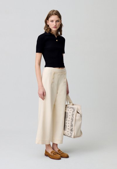 Haut noir à manches courtes à côtes, pantalon large crème avec des boutons, mocassins marron et un sac beige avec des détails paisley.