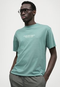 Tricou mint, realizat din bumbac moale, cu mânecă scurtă și guler rotund. Logo imprimat în alb, centrat. Design și formă simple.