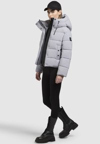 Graue Pufferjacke mit hohem Kragen, Reißverschluss und Druckknopfverschlüssen sowie schwarzen Akzenten. Kombiniert mit schwarzen Leggings und klobigen, schwarzen Schnürstiefeln.