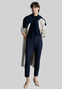 Beige Trenchcoat, dunkelblauer Pullover am Hals gebunden, taillierte dunkelblaue Hose und nudefarbene High Heels, präsentiert vor einem neutralen Hintergrund.