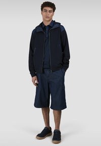 Giacca a vento blu con cappuccio, abbinata a una polo blu e pantaloni cargo fino al ginocchio. Sneakers scure completano l'outfit.