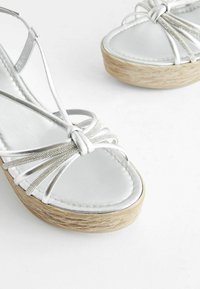 Stříbrné sandály na tenkých páscích s texturovanou espadrilovou wedge podrážkou, s hladkým lesklým svrškem a dekoračními uvázanými pásky.