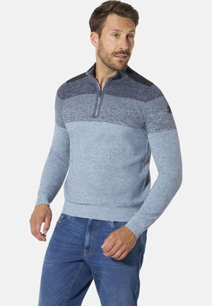 Mann trägt einen blau-grauen Strickpullover mit Viertelreißverschluss und Schulterpatches, kombiniert mit blauen Jeans, steht vor einem schlichten Hintergrund.