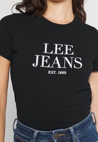 Camiseta de algodón negra con "LEE JEANS" y "EST. 1889" impresos en blanco. Presenta un escote redondo y un ajuste cómodo. Combinada con jeans de mezclilla azul.