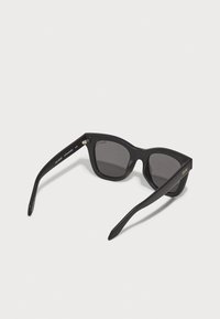Lunettes de soleil noires avec une finition mate, forme angulaire, verres sombres et accent doré sur la branche, présentant un design robuste et des charnières polies.