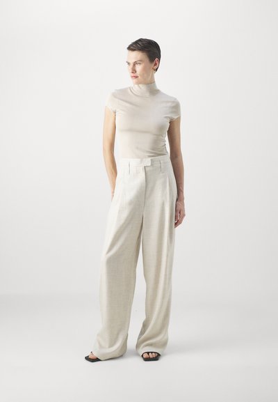 Μπεζ εφαρμοστό τοπ με τρουακάρ γιακά, συνδυασμένο με ελαφριά λινά παντελόνια. Τα παντελόνια έχουν φαρδιά πόδια και ψηλή μέση. Το μοντέλο φορά μινιμαλιστικά μαύρα σανδάλια.