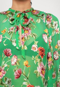 Bluză florală verde, cu un guler înalt și detaliu cu panglică, din material transparent, cu modele de flori multicolore pe față.