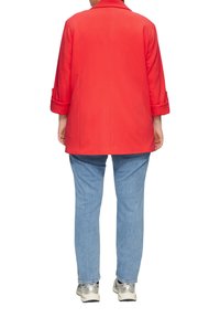 Personne portant une veste rouge, un jean bleu clair et des baskets blanches, se tenant de dos sur un fond blanc.