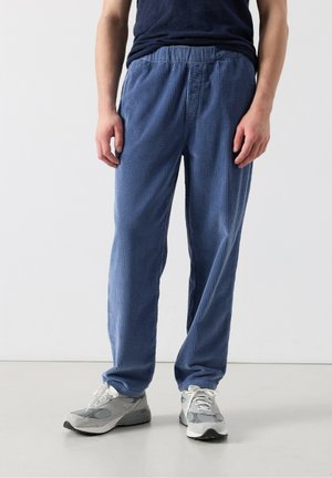 Muž oblečený v modrých kordových nohaviciach, námorníckej košeli a svetlosivých teniskách, stojaci pred jednofarebným bielym pozadím.