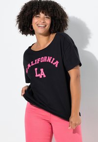 Mujer sonriente con cabello rizado, vistiendo una camiseta negra que dice "California LA" y pantalones rosas, de pie contra una pared blanca.