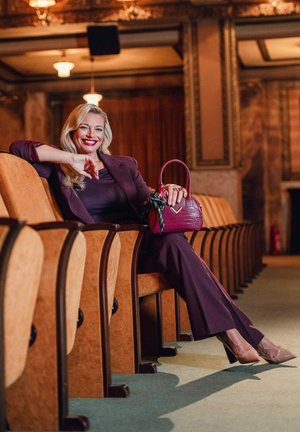 Švestkově zbarvený oblek, přiléhavý top a zvonové kalhoty. Drží texturovanou vínově červenou kabelku s květinovými detaily. Béžové podpatky, sedí v divadle.